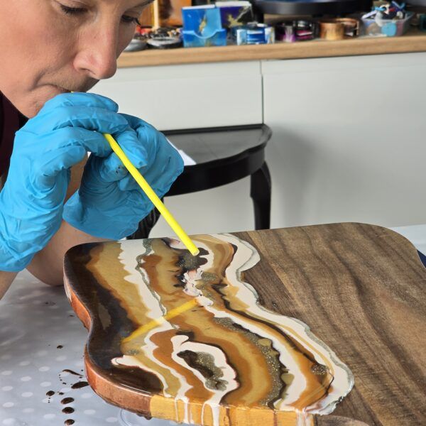 Lady with blue gloves blowing paint on a cheese board via a yellow straw.